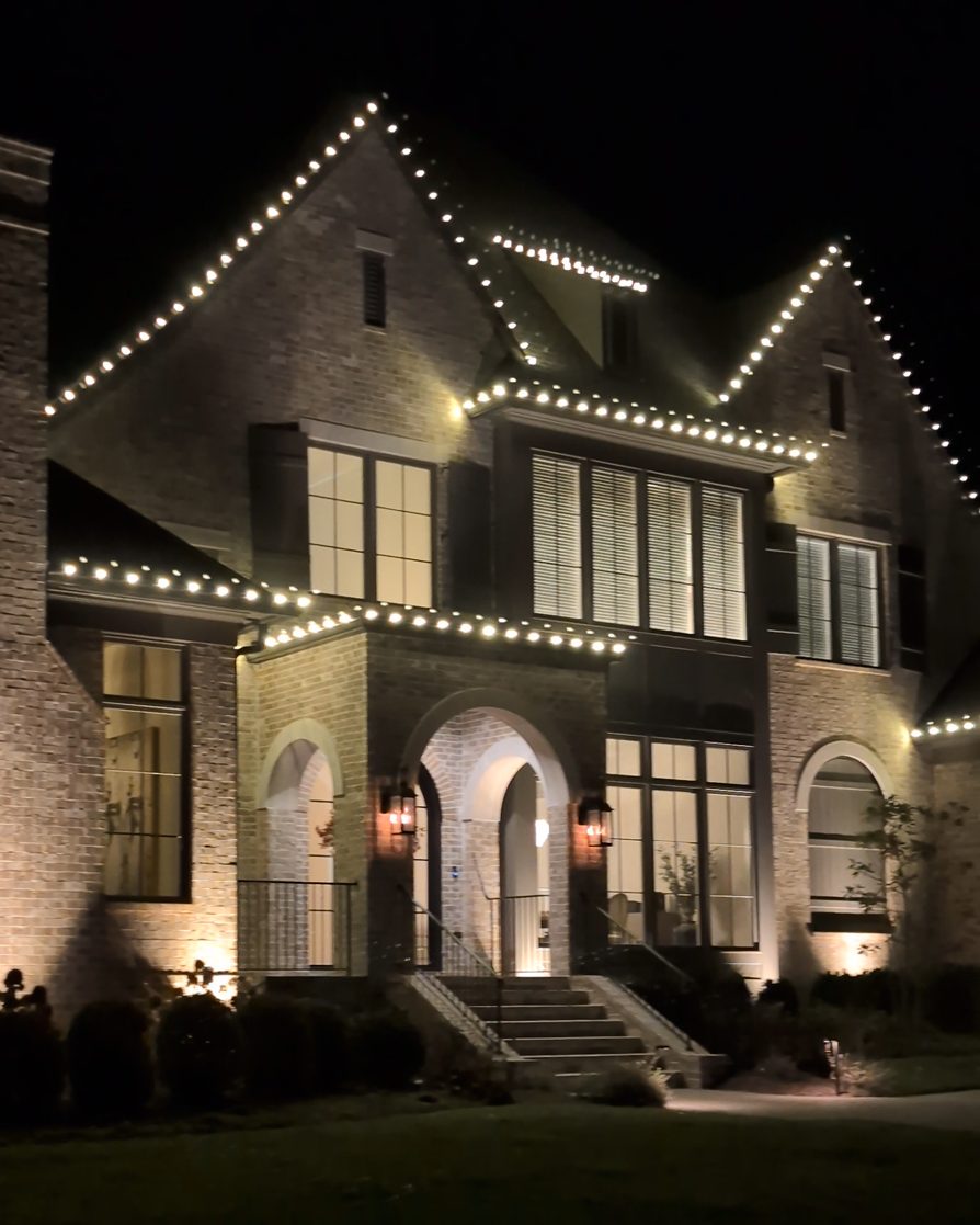 The same brick home at night with warm white lights along every roofline