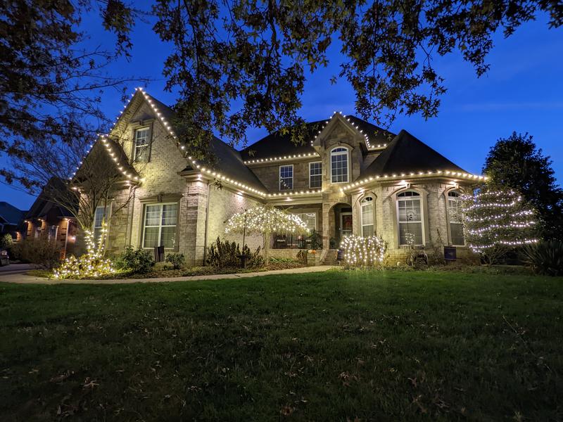 Large brick home at dusk with lit trees and white roofline lights