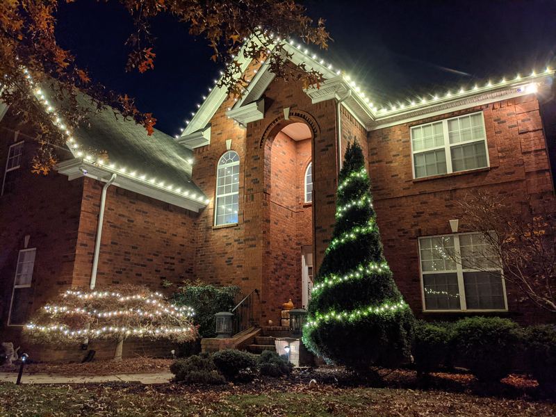 Brick home with white roofline lights, a lit tree and a wrapped evergreen