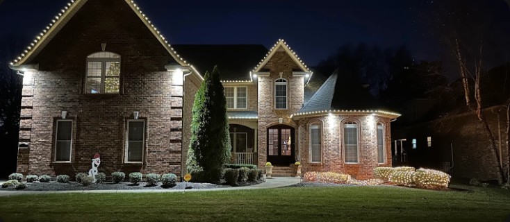 Brick home with white lights along every roofline and glowing shrubs