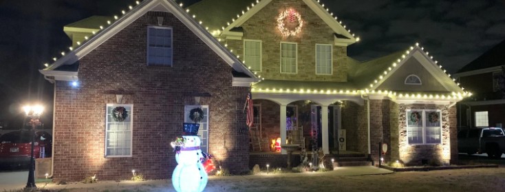 Brick home with white lights on every peak, a lit wreath and a snowman out front